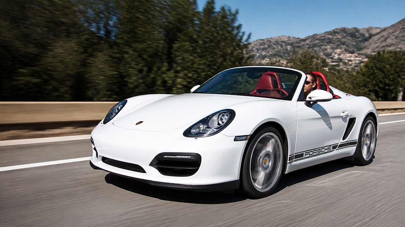 Carro esportivo conversível branco Porsche dirigindo em estrada com paisagem montanhosa ao fundo.
