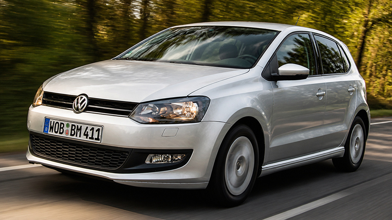 Carro Volkswagen prata em movimento em estrada rodeada por árvores com placas europeias.