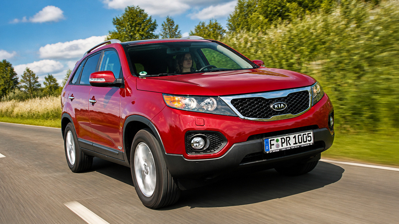 Carro SUV vermelho Kia em movimento em estrada cercada por árvores e céu azul com nuvens.