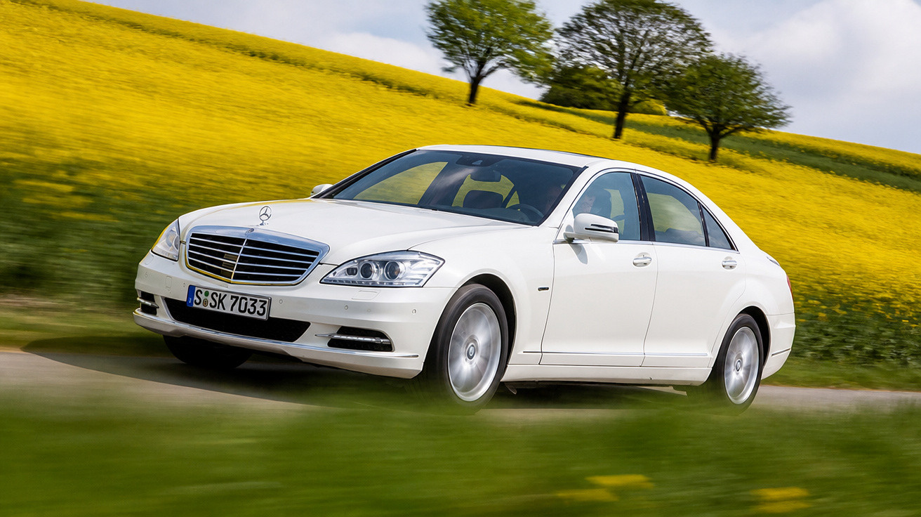Carro branco Mercedes-Benz em movimento em estrada cercada por campo com flores amarelas e árvores ao fundo.