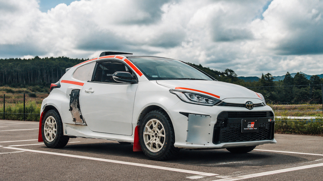 Carro esportivo branco com detalhes vermelhos, rodas off-road e para-lamas danificados estacionado em área externa.