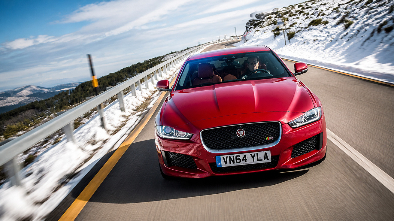 Carro esportivo vermelho Jaguar dirigindo em estrada de montanha com neve nas laterais e céu azul.