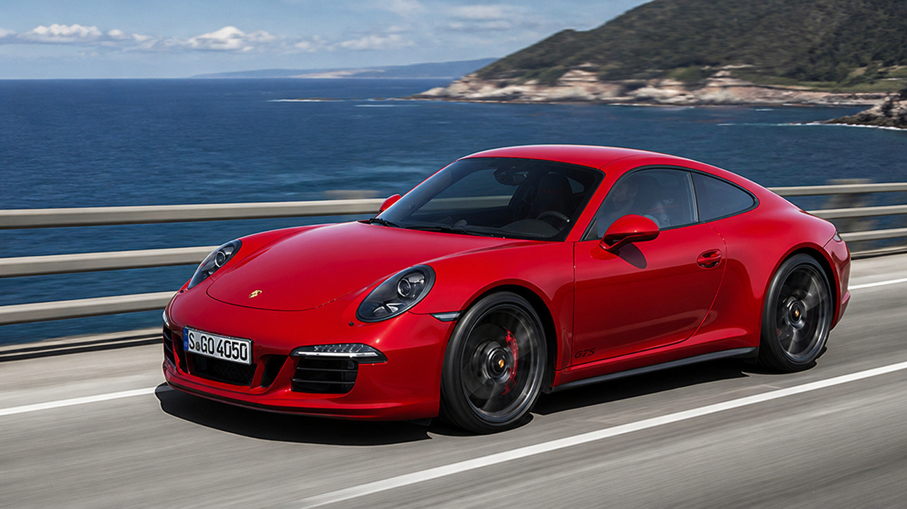 Carro esportivo vermelho Porsche 911 GT3 em alta velocidade em estrada à beira-mar com montanha ao fundo.
