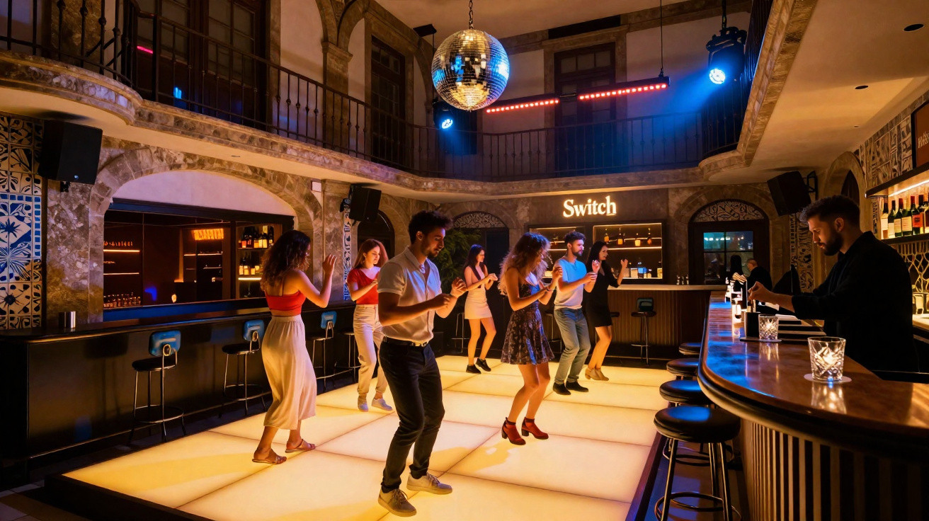 Pessoas dançando em pista iluminada em bar com decoração vintage e DJ ao balcão.