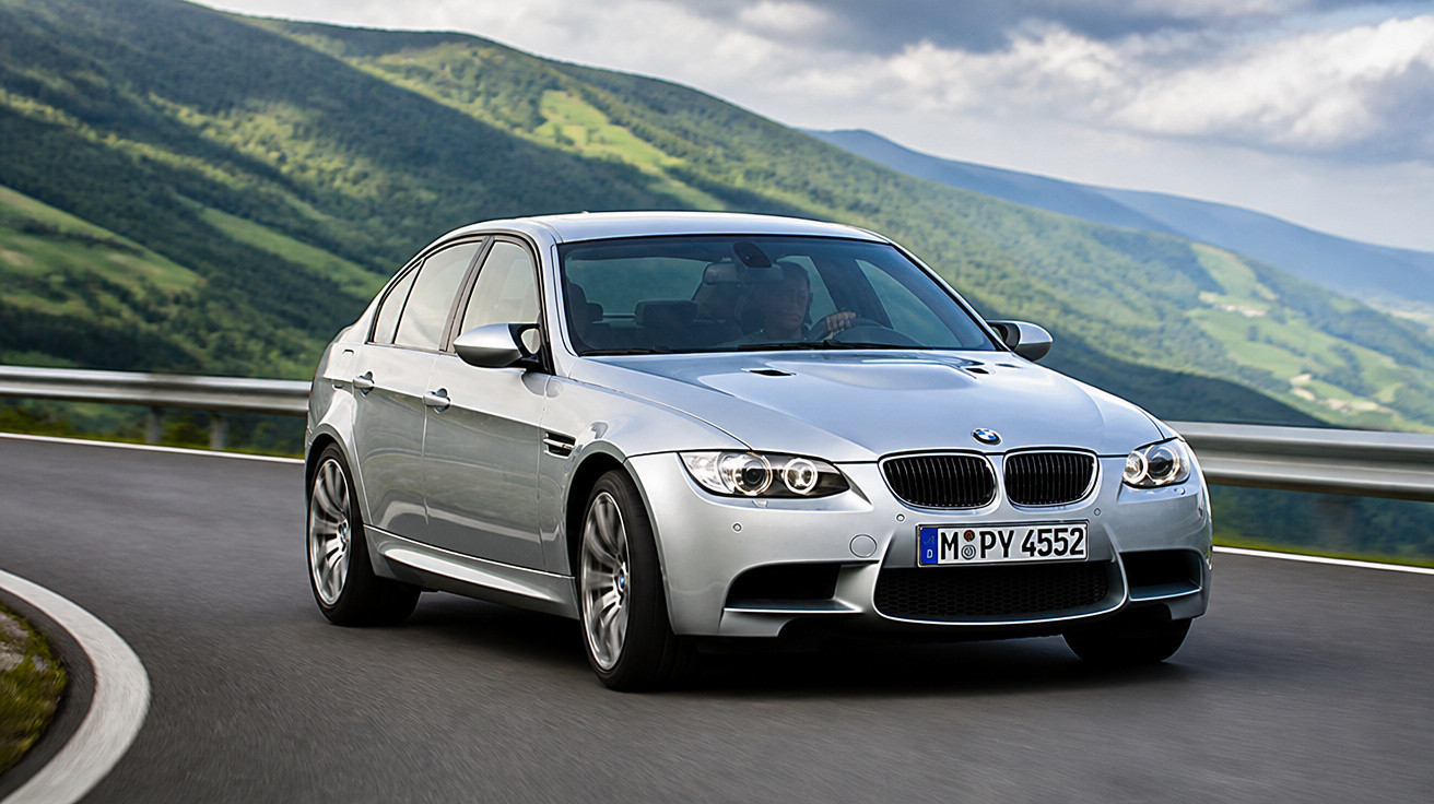 Carro prata BMW M3 dirigindo em estrada sinuosa com montanhas verdes ao fundo sob céu nublado.