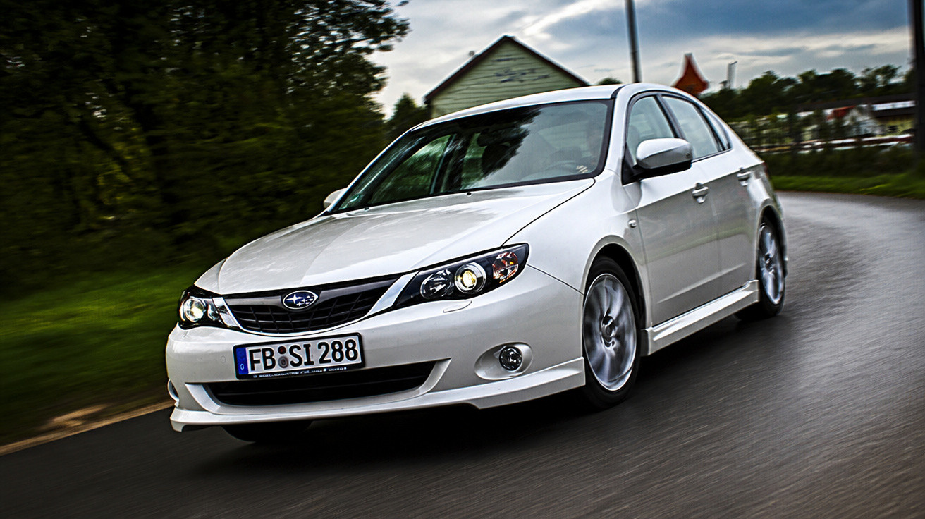 Carro branco Subaru sedan em movimento em estrada curva com árvores e casas ao fundo.