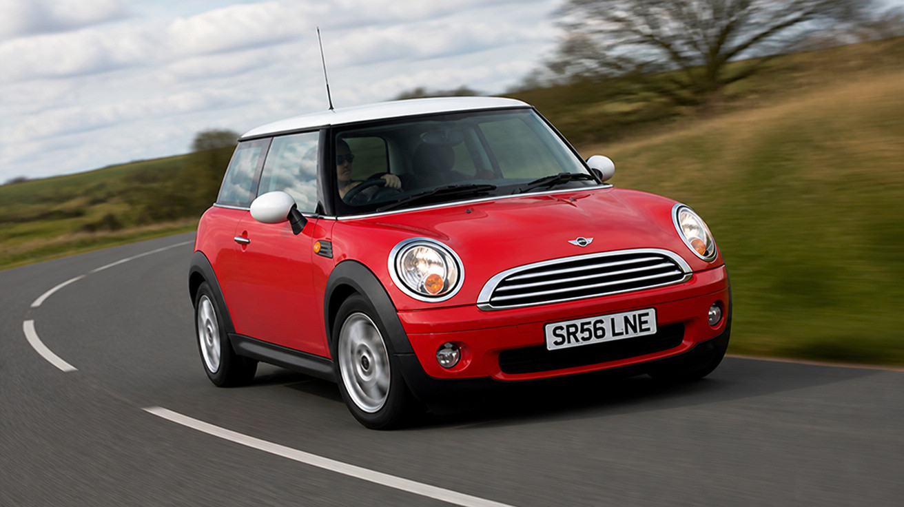 Carro Mini Cooper vermelho com teto branco dirigindo em estrada curva cercada por árvore e campo.