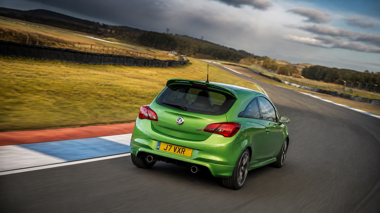 Carro verde esportivo em curva de pista de corrida com céu nublado ao fundo.