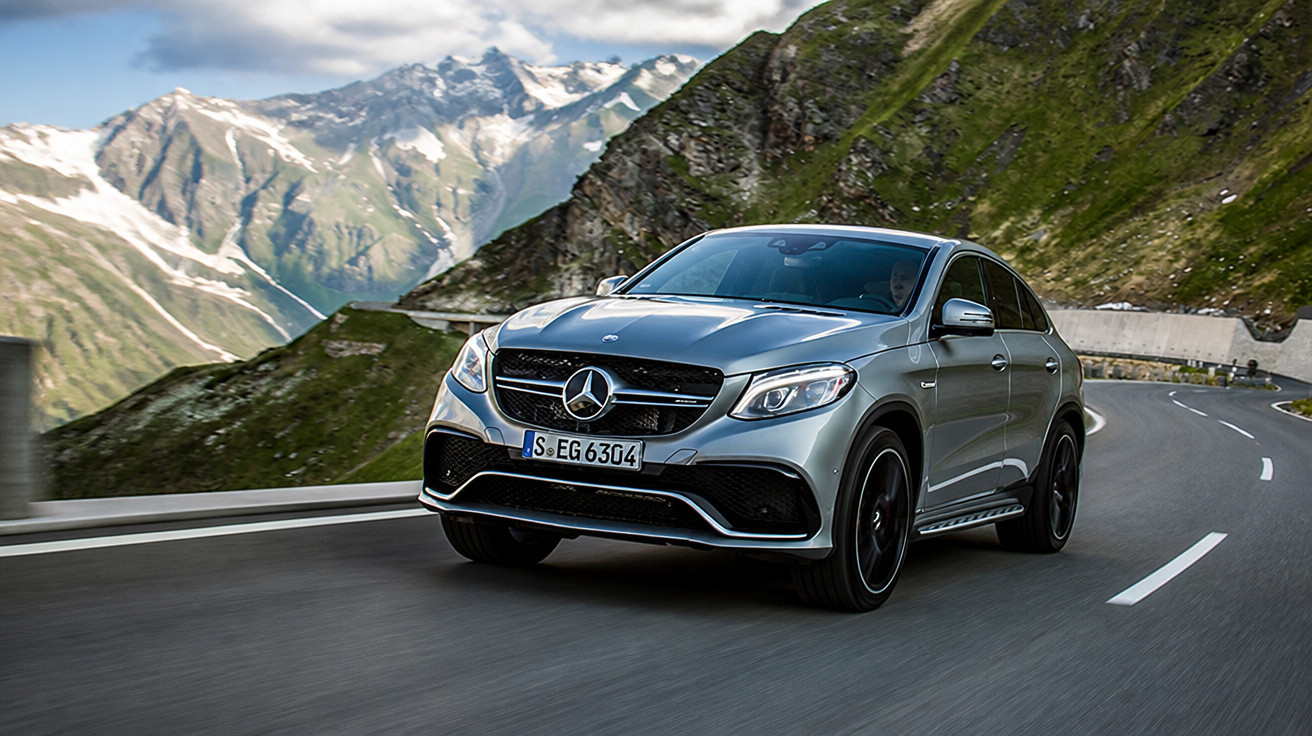 Carro Mercedes-Benz prata trafegando em estrada sinuosa cercada por montanhas verdes e com picos nevados ao fundo.