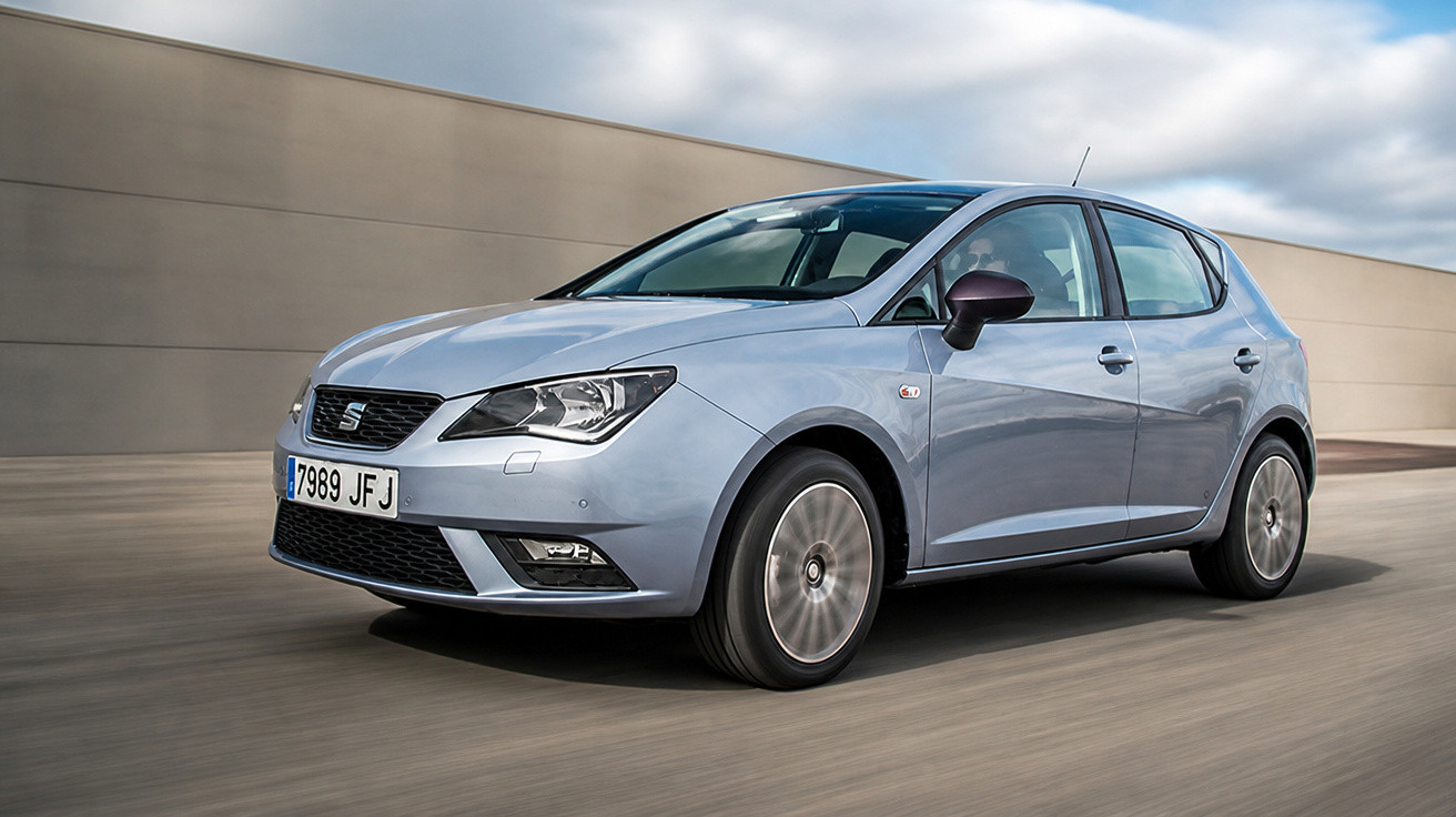 Carro hatchback cinza em movimento com fundo urbano desfocado em dia nublado.