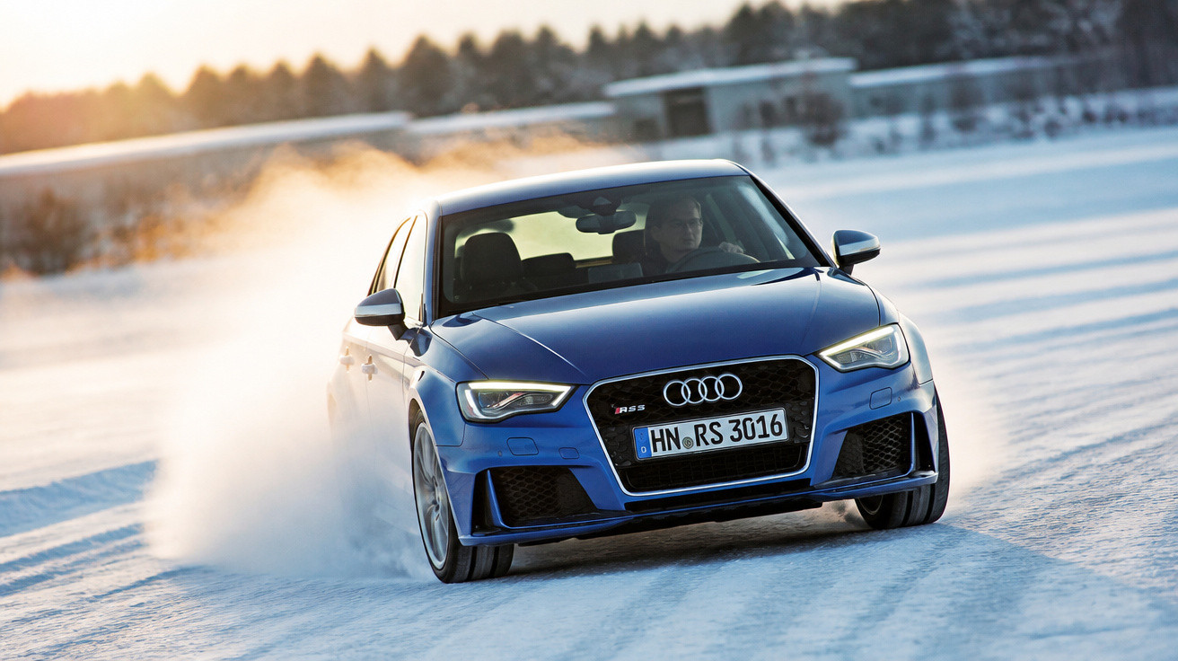 Carro esportivo azul Audi RS 3 dirigindo em estrada coberta de neve com céu ao entardecer.