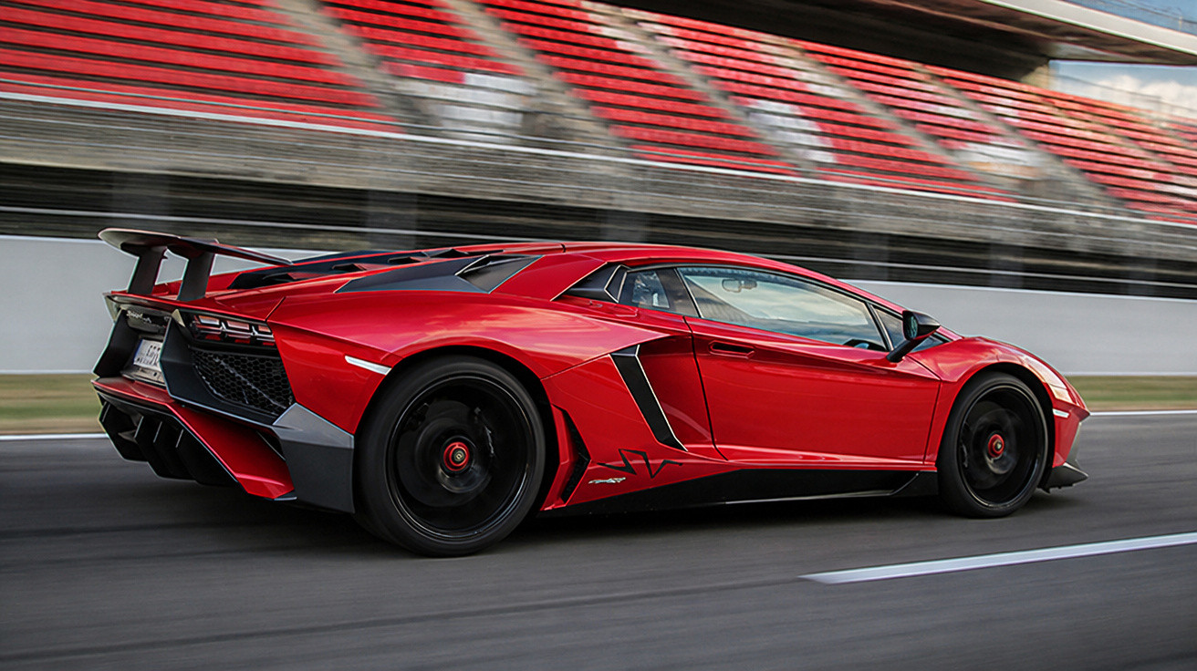 Carro esportivo vermelho em alta velocidade em pista de corrida com arquibancadas ao fundo.