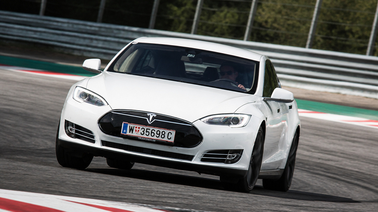 Carro elétrico Tesla branco em curva fechada em pista de corrida com cerca metálica ao fundo.