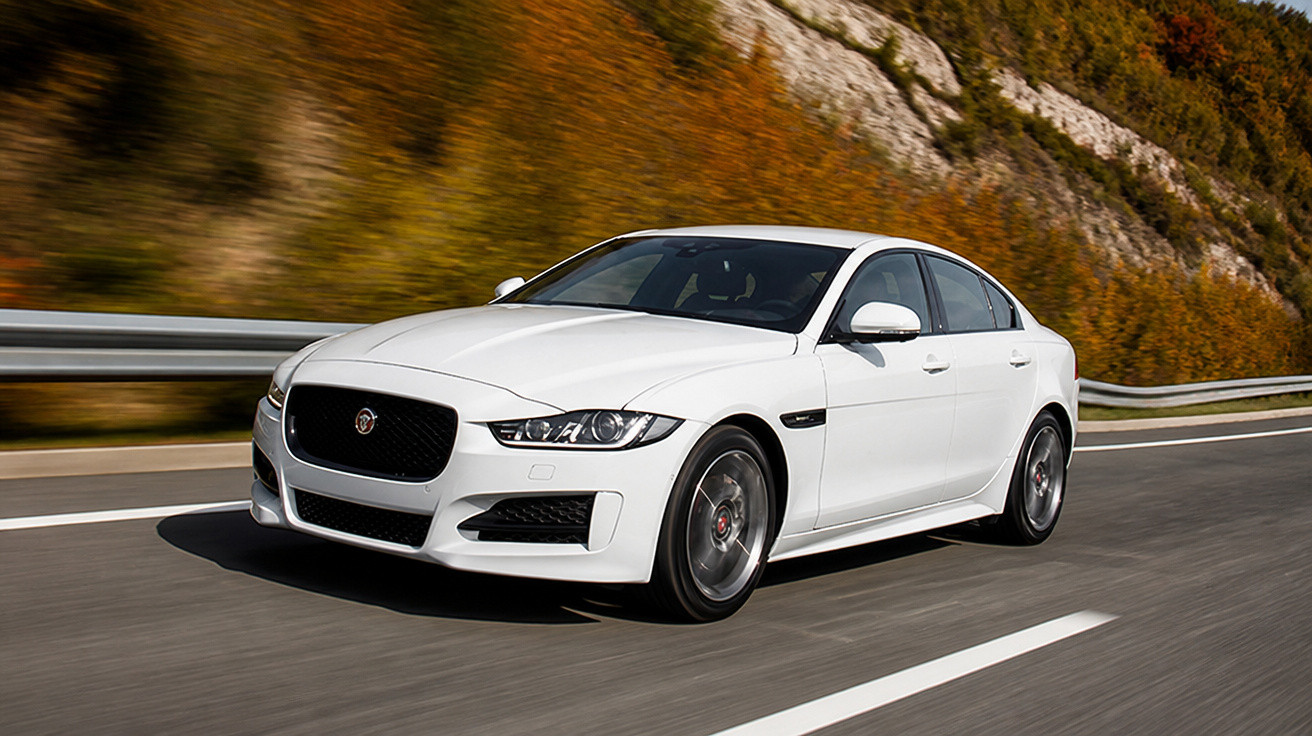 Carro branco esportivo sedan Jaguar circulando em estrada asfaltada com vegetação ao fundo.