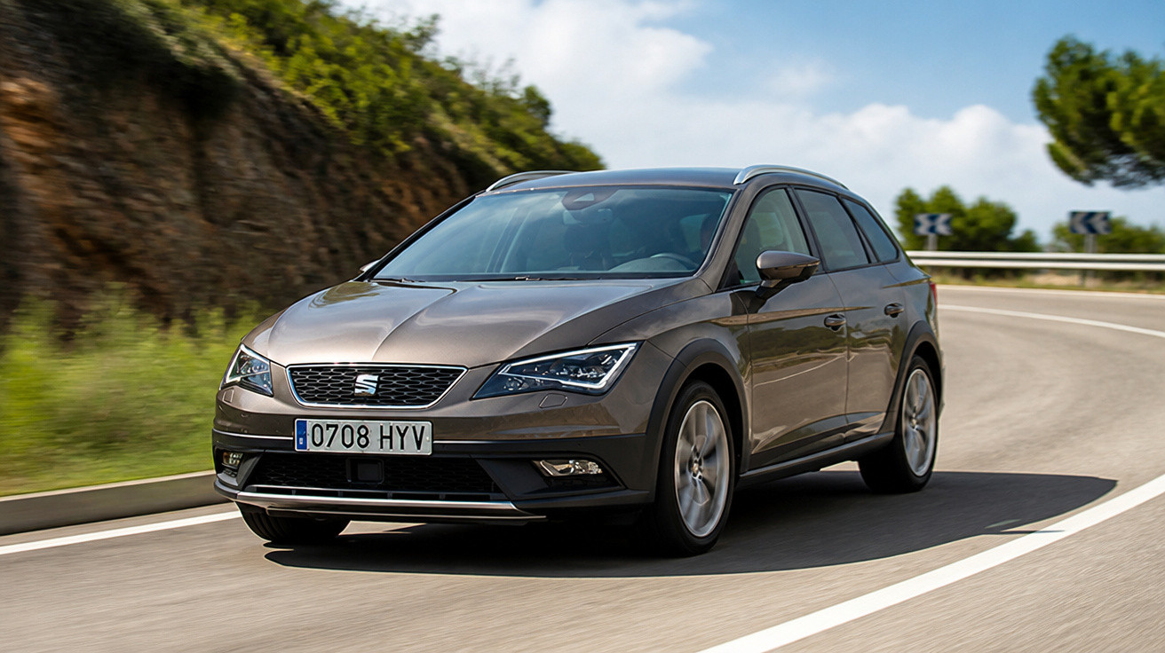 Carro cinza modelo SEAT Cruze trafegando em curva de estrada asfaltada sob céu azul com nuvens.