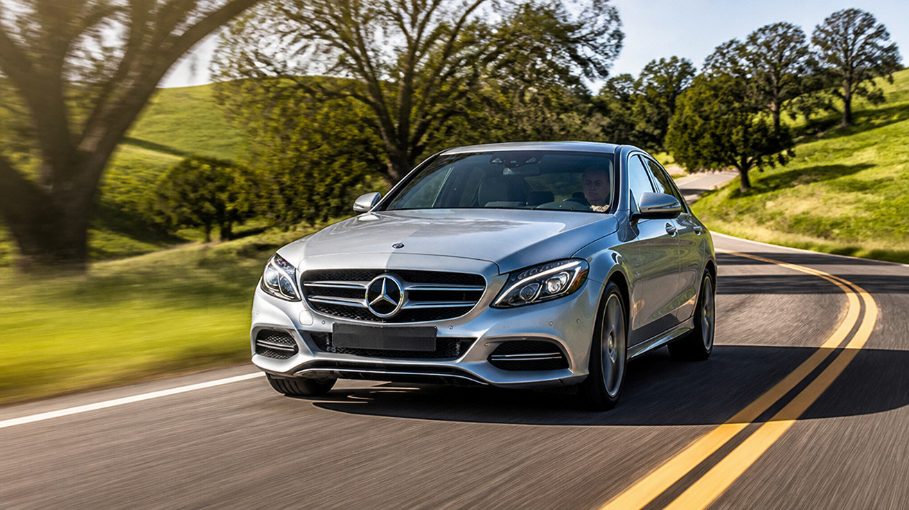 Carro prata Mercedes-Benz modelo sedan dirigindo em estrada asfaltada com árvores e colinas verdes ao redor.