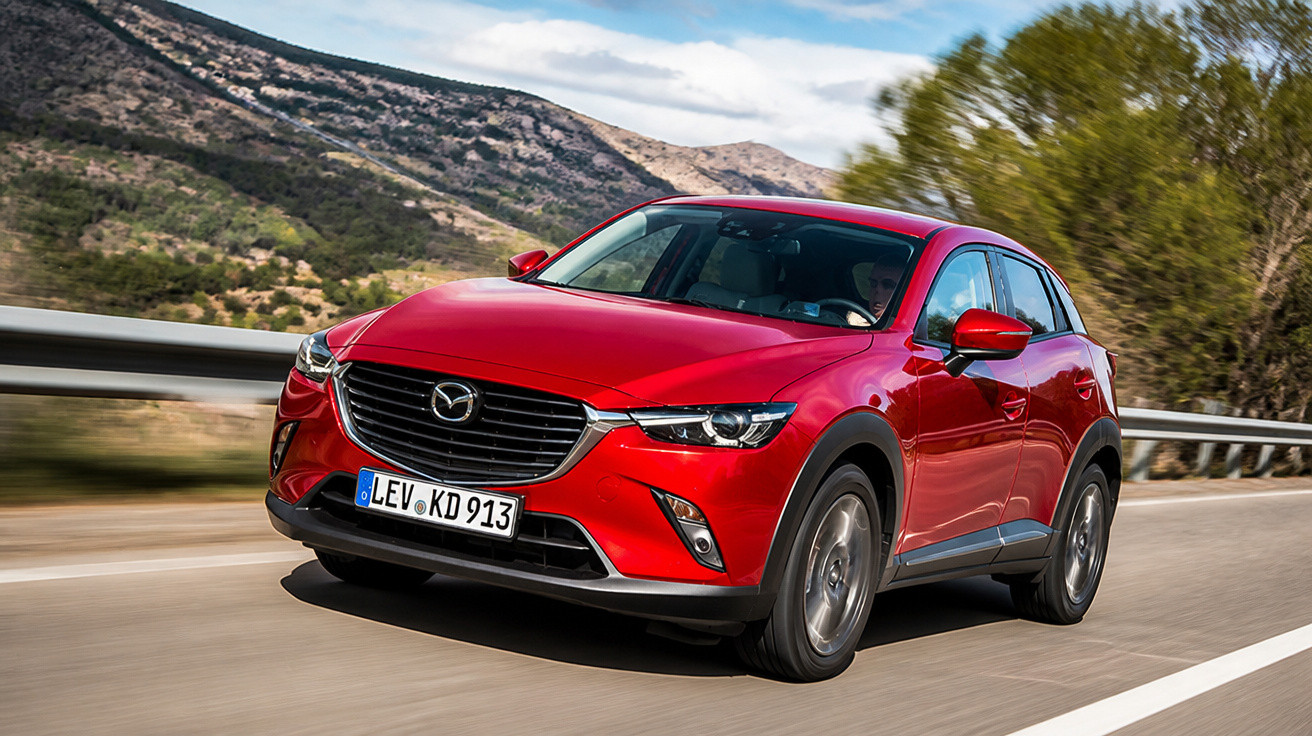 Carro vermelho Mazda CX-3 em movimento numa estrada cercada por árvores e montanhas.