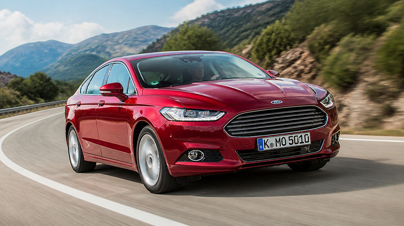 Carro vermelho Ford Fusion em movimento em estrada sinuosa com montanhas ao fundo.