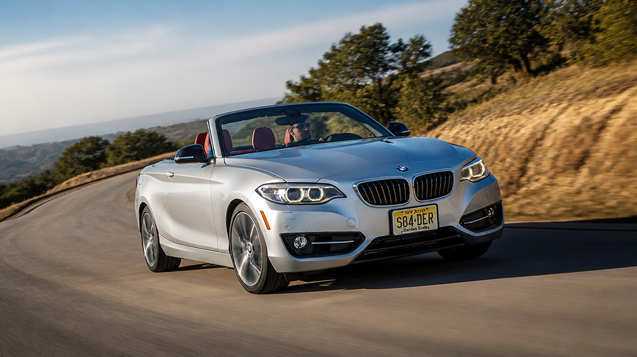Carro conversível prata BMW dirigindo em estrada sinuosa com paisagem de árvores e morros ao fundo.