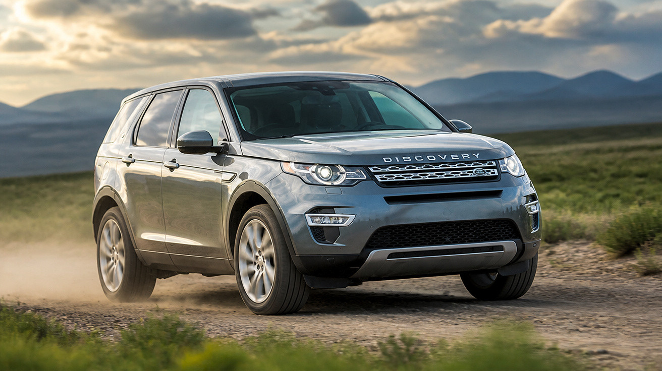 SUV cinza Land Rover Discovery em estrada de terra com montanhas ao fundo e céu nublado.