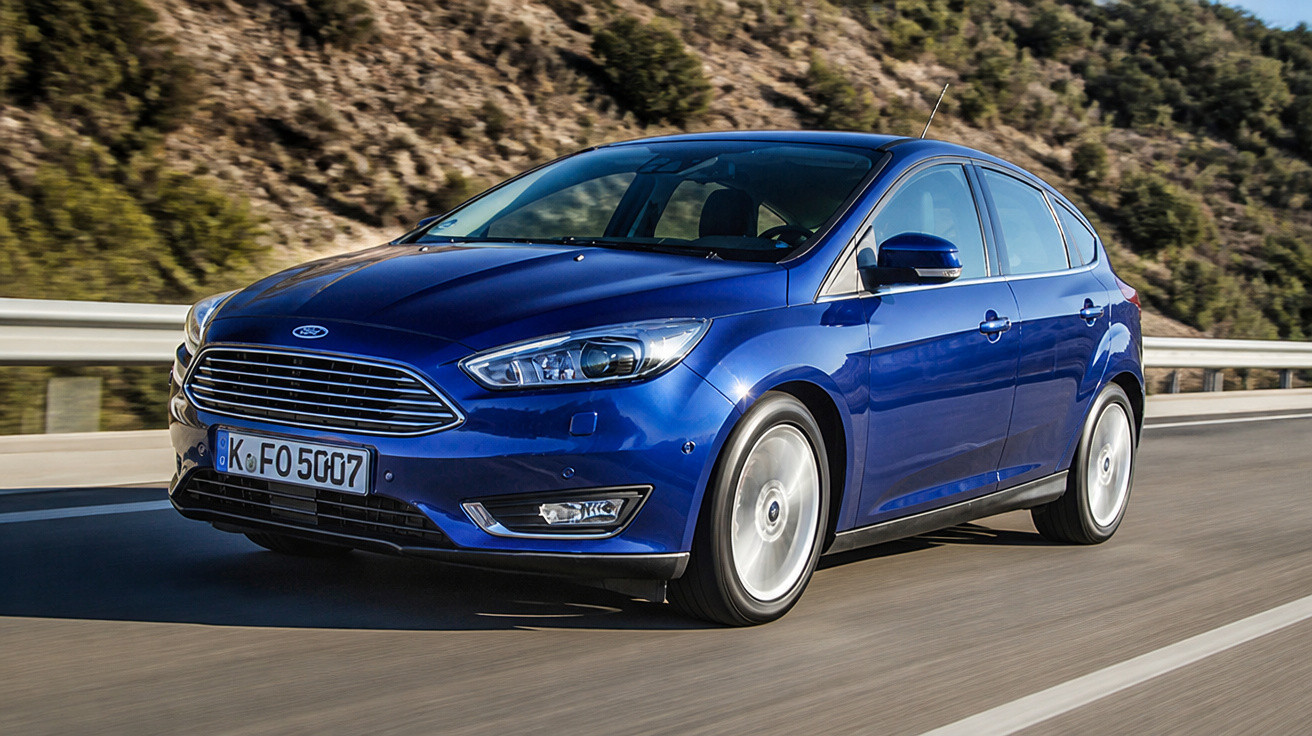 Carro hatchback azul em movimento em estrada com paisagem montanhosa ao fundo.