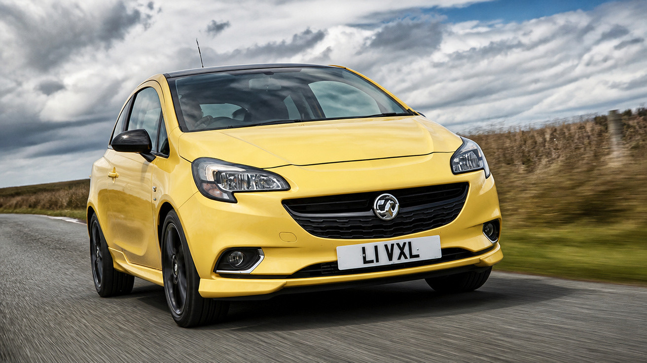Carro compacto amarelo modelo Vauxhall circulando em estrada com céu nublado e vegetação ao redor.