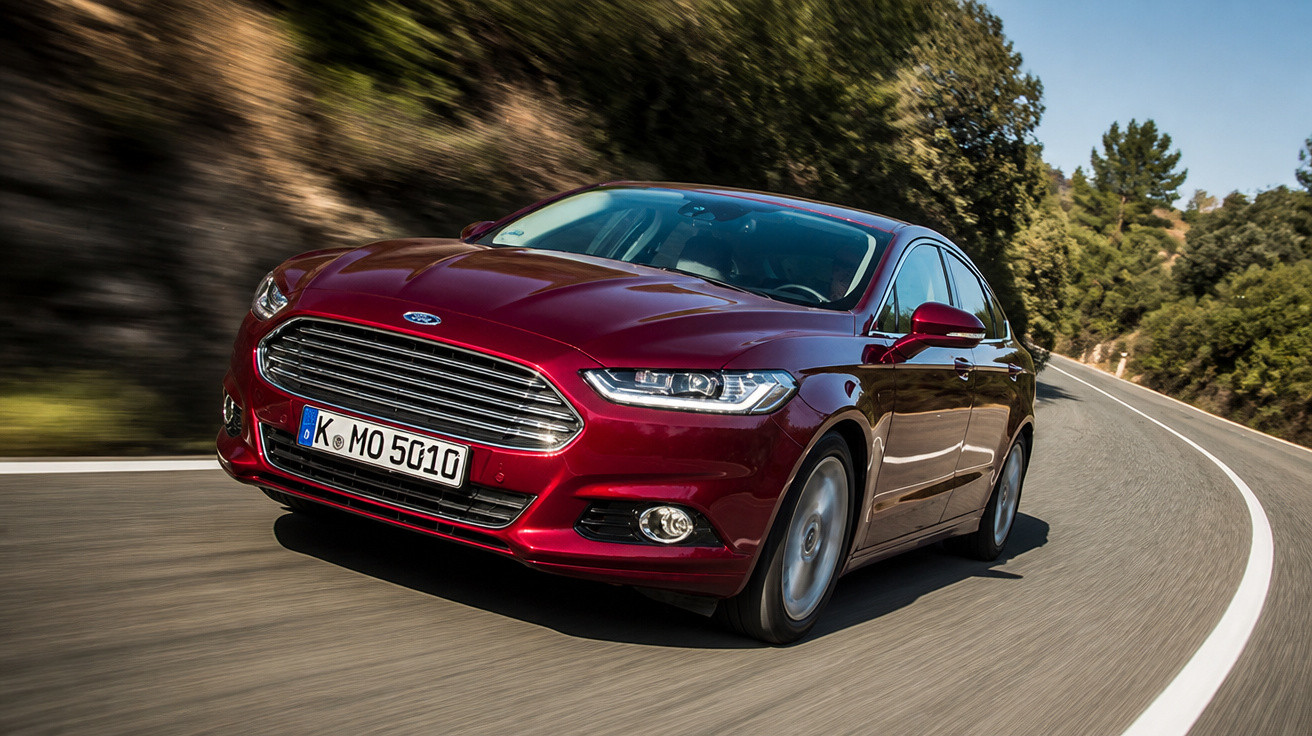 Carro Ford vermelho em movimento em estrada sinuosa cercada por árvores e vegetação.
