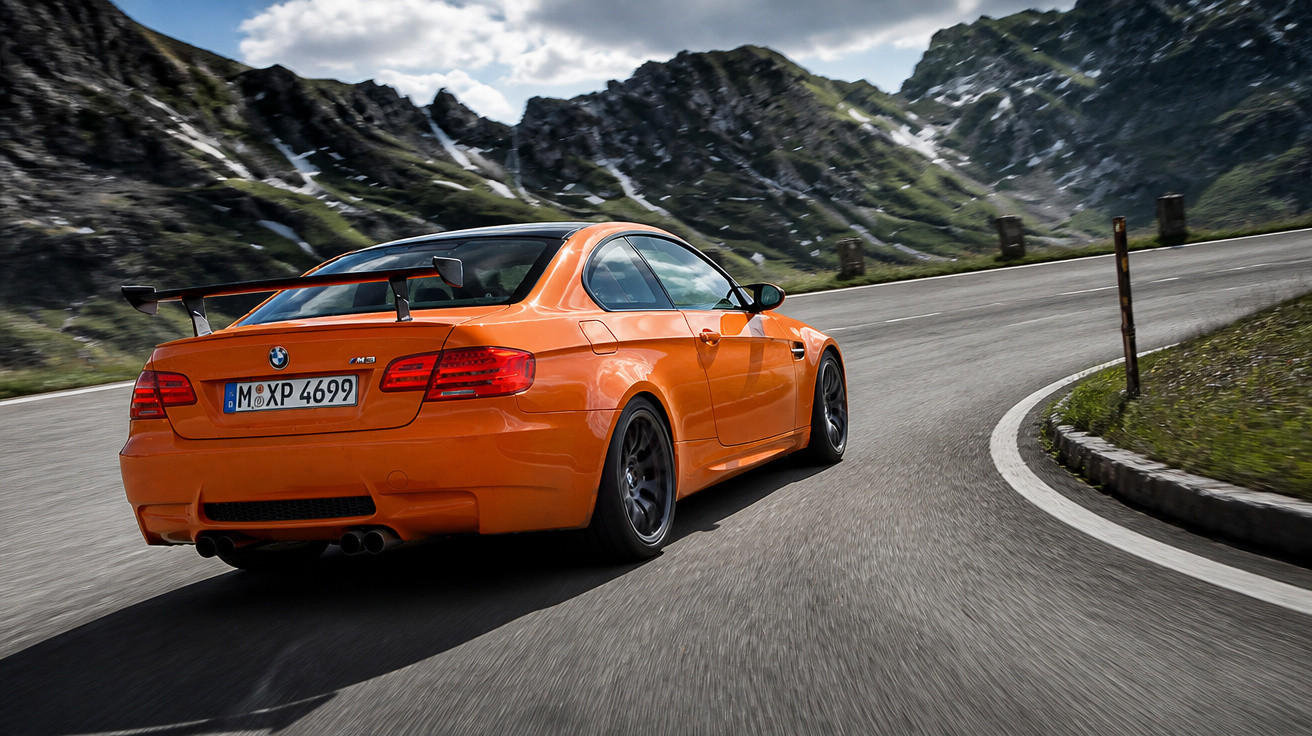Carro esportivo laranja BMW M3 em curva de estrada sinuosa com montanhas ao fundo.