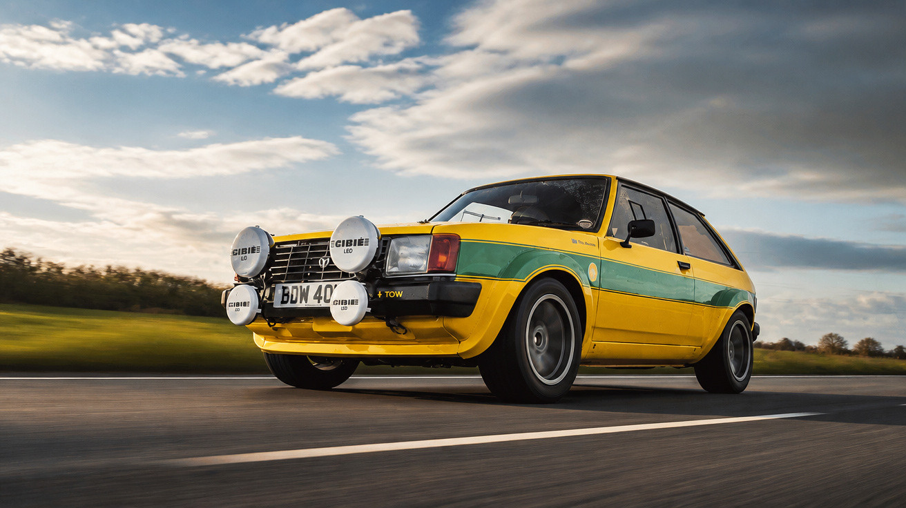 Carro hatch amarelo com faixa verde, faróis extras, em estrada sob céu nublado ao entardecer.