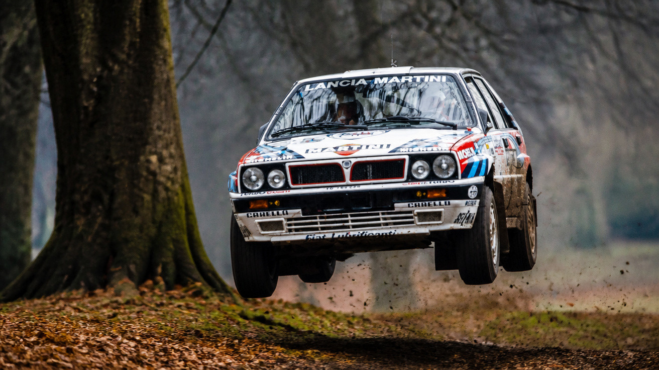 Carro de rali Lancia Delta branco com patrocínios saltando em estrada de terra cercada por árvores.