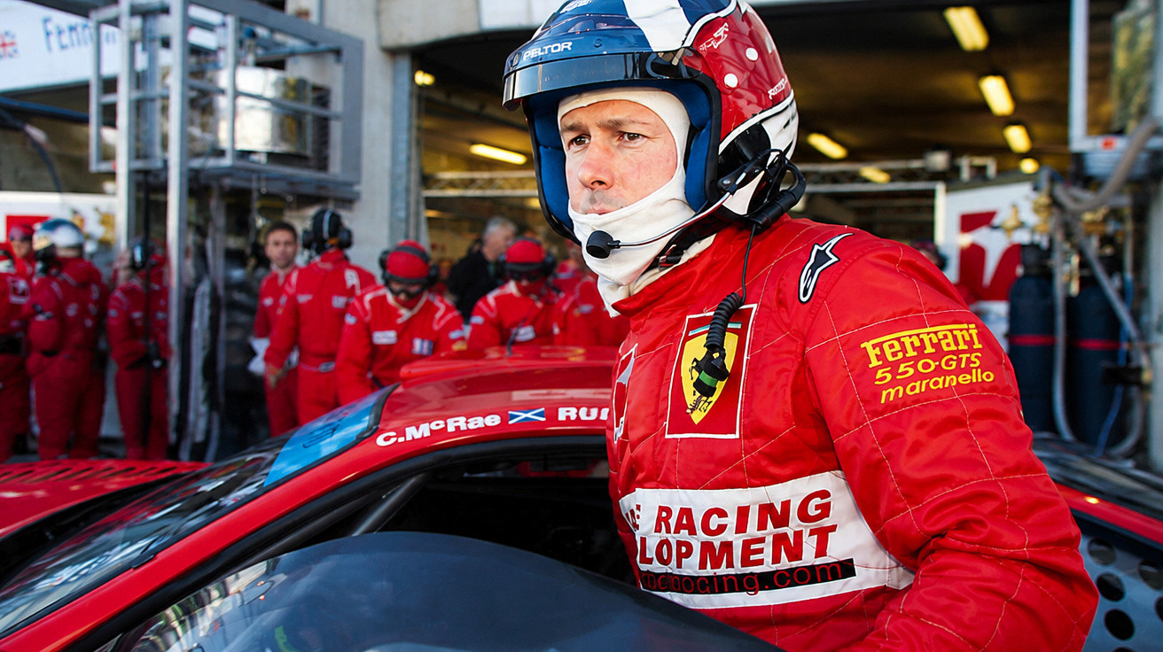 Piloto de corrida com macacão vermelho e capacete azul ao lado de carro esportivo Ferrari na garagem.