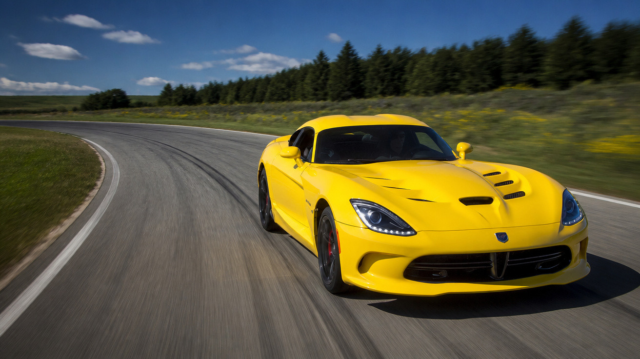 Carro esportivo amarelo acelerando em pista curva com céu azul e árvores ao fundo.