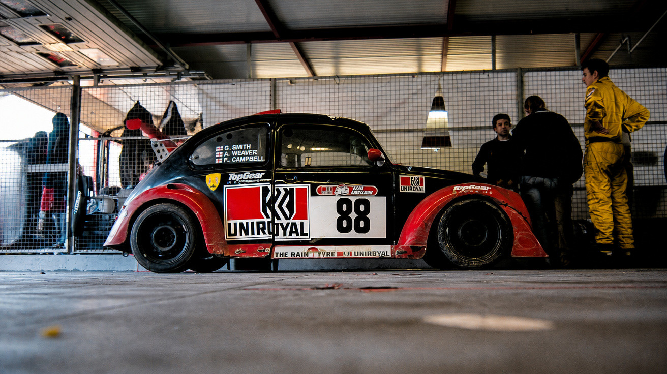 Carro de corrida modelo Fusca com pintura vermelha e preta, número 88, estacionado em garagem com três pessoas ao fundo.
