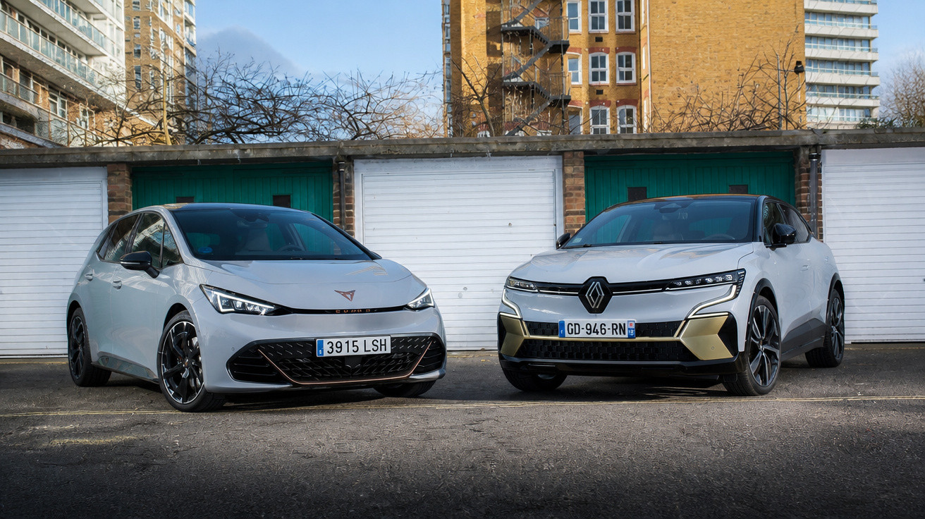 Dois carros elétricos brancos estacionados em frente a garagens com edifícios ao fundo em área urbana.