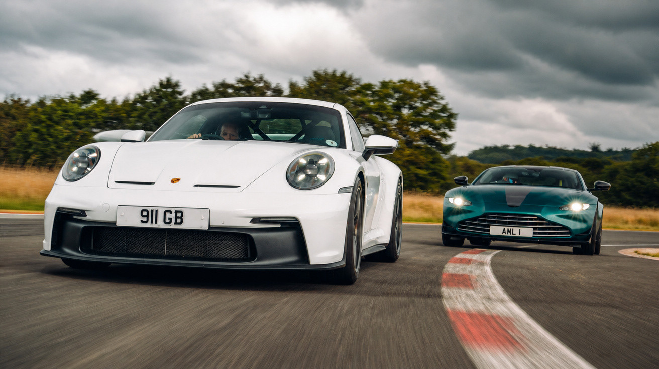 Dois carros esportivos em alta velocidade em pista de corrida com céu nublado ao fundo.
