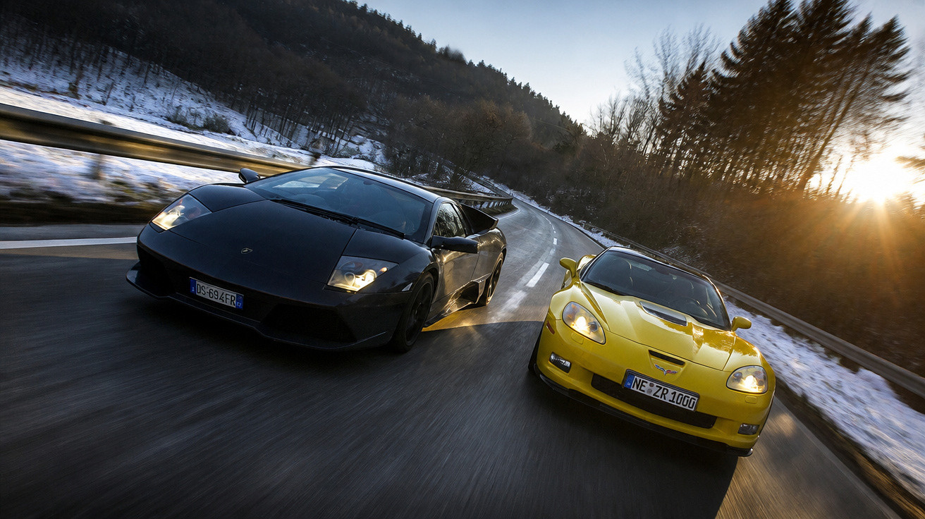 Dois carros esportivos, um preto e um amarelo, acelerando em estrada sinuosa com neve nas laterais ao pôr do sol.