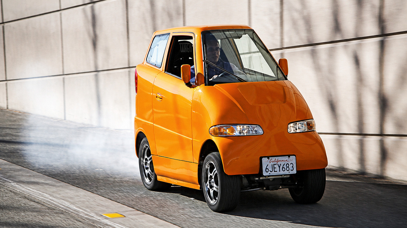 Carro compacto e laranja com três rodas dirigindo em rua urbana ensolarada.