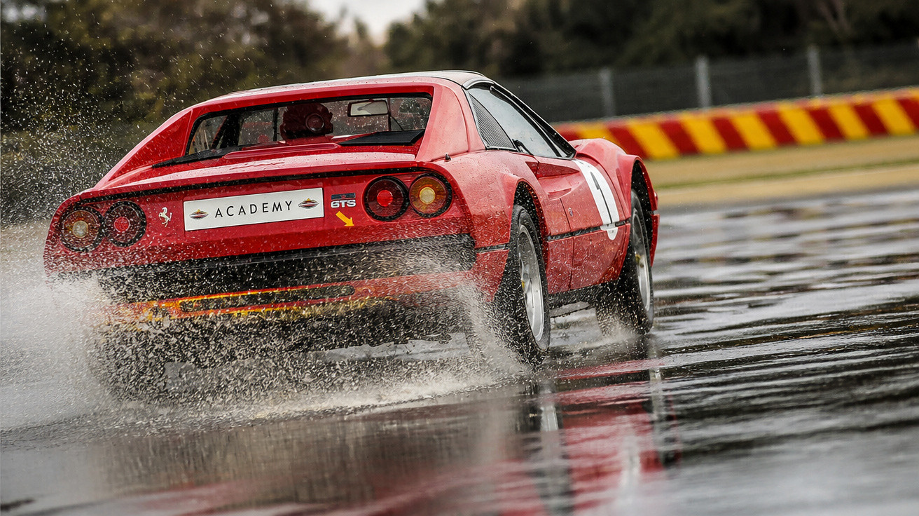 Carro esportivo vermelho acelerando em pista molhada com respingos de água ao redor.