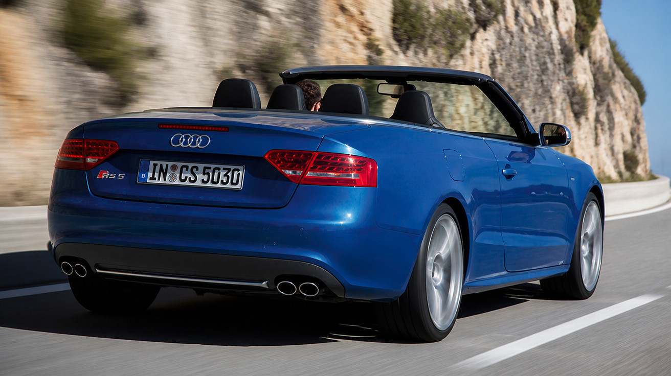 Carro conversível azul Audi RS5 sendo dirigido em estrada com penhasco ao lado em dia ensolarado.