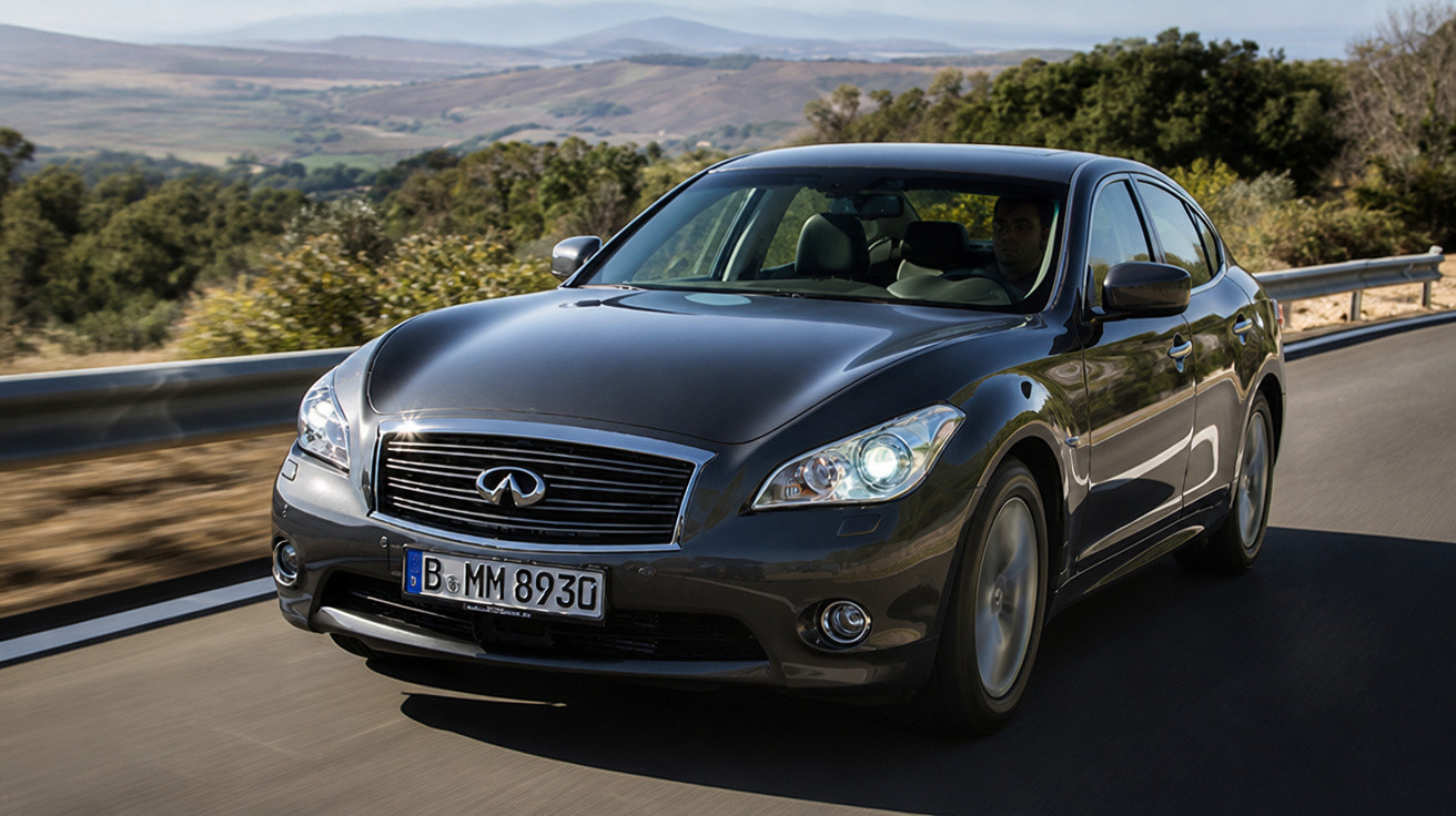 Carro preto da marca Infiniti trafegando em estrada com paisagem montanhosa ao fundo.