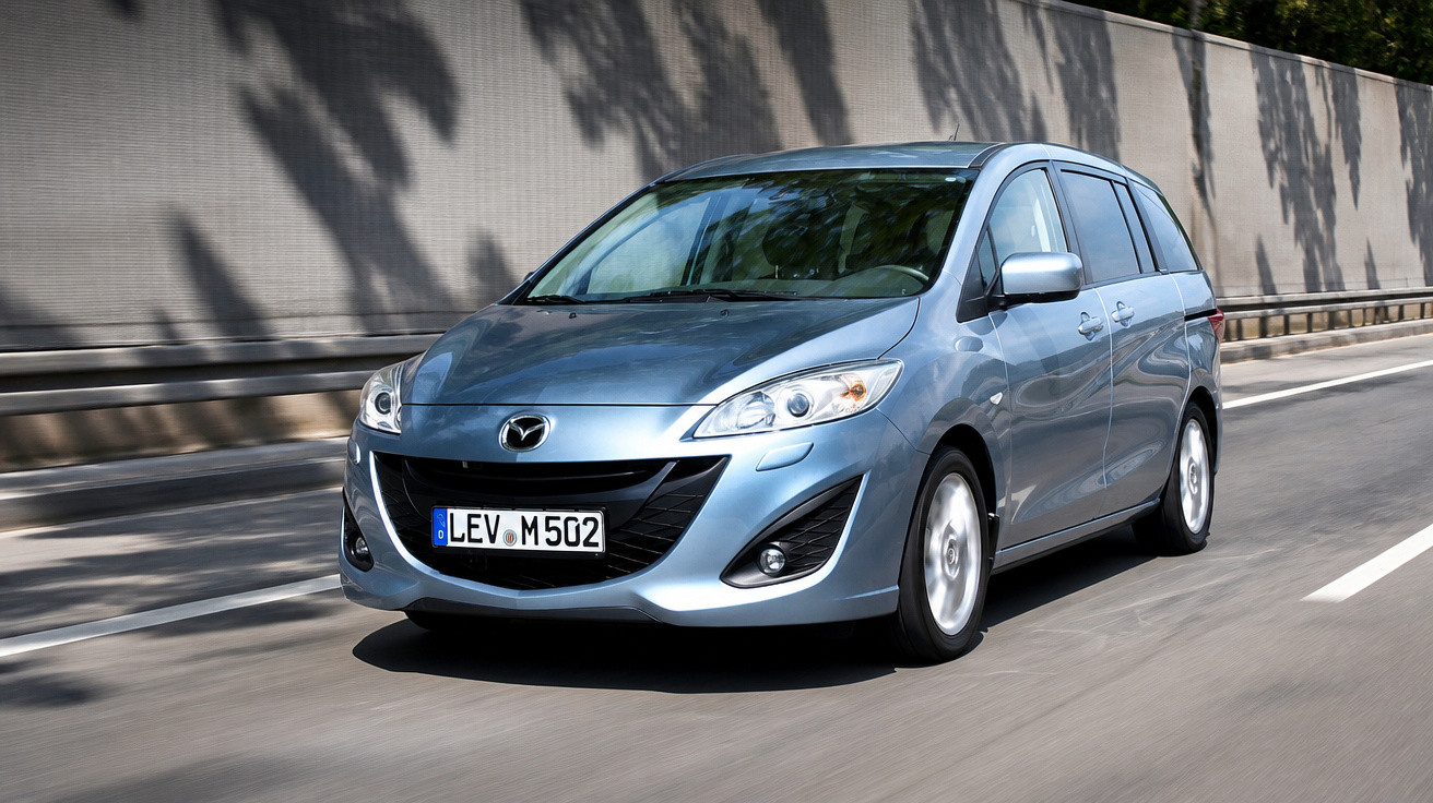 Carro cinza modelo Mazda 5 em movimento em uma estrada urbana com muro lateral.