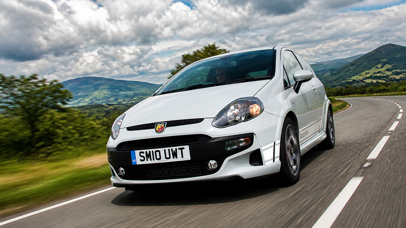 Carro esportivo branco Abarth em estrada sinuosa com montanhas e céu nublado ao fundo.