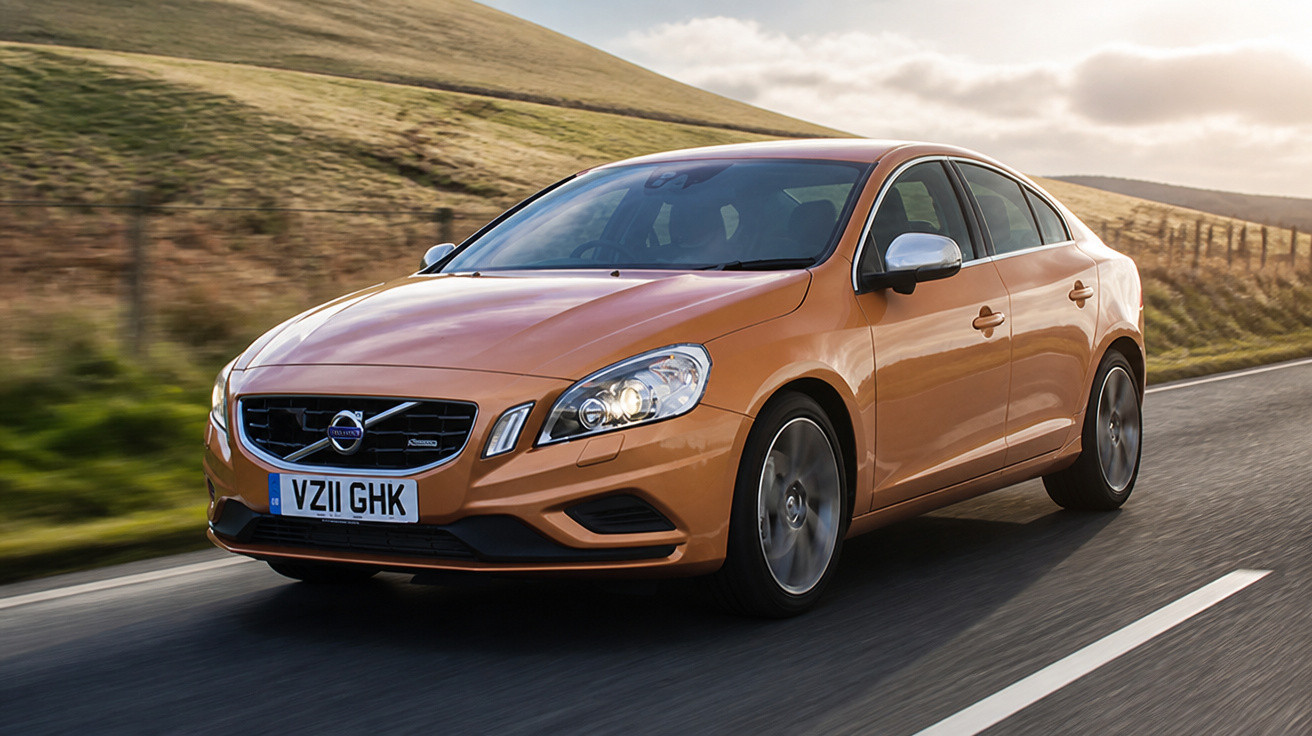 Carro sedan laranja Volvo em movimento na estrada com colinas ao fundo e céu nublado.
