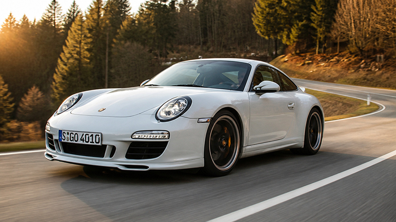 Carro esportivo branco Porsche 911 em curva de estrada cercada por árvores ao pôr do sol.
