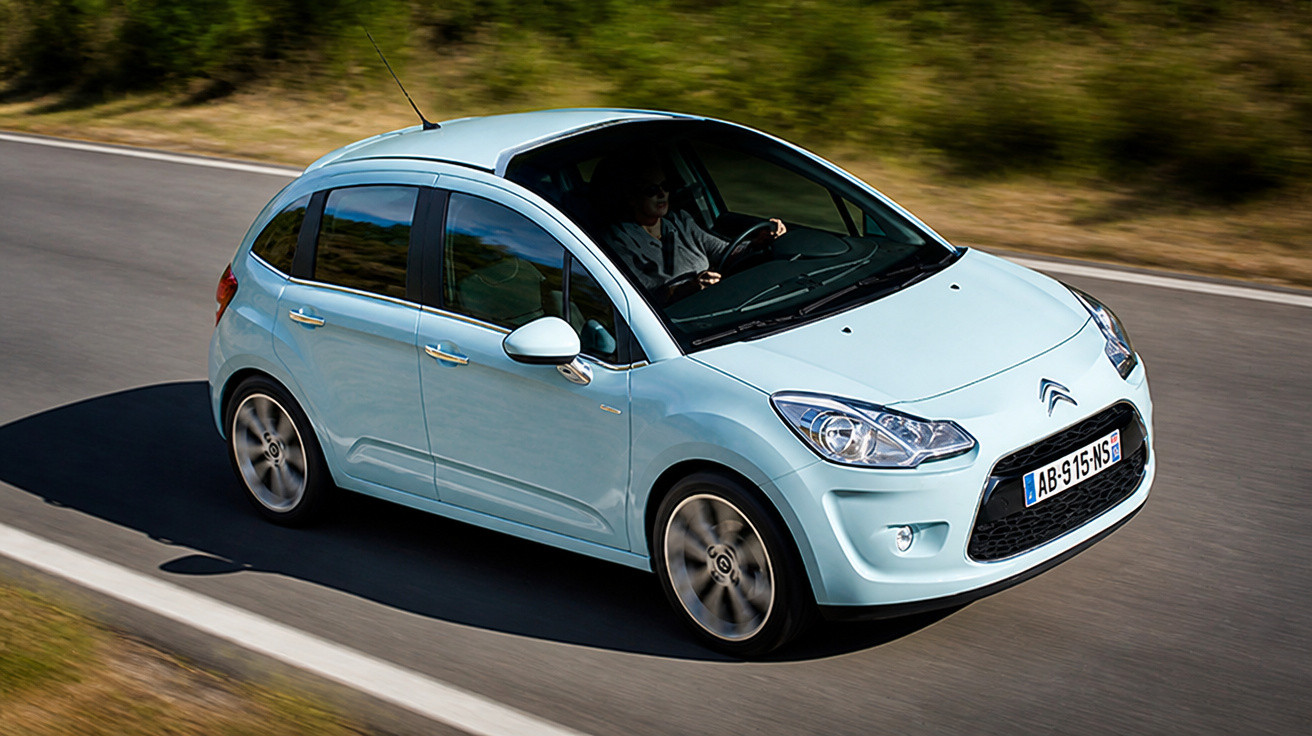 Carro hatchback azul claro em movimento em estrada asfaltada com motorista ao volante.