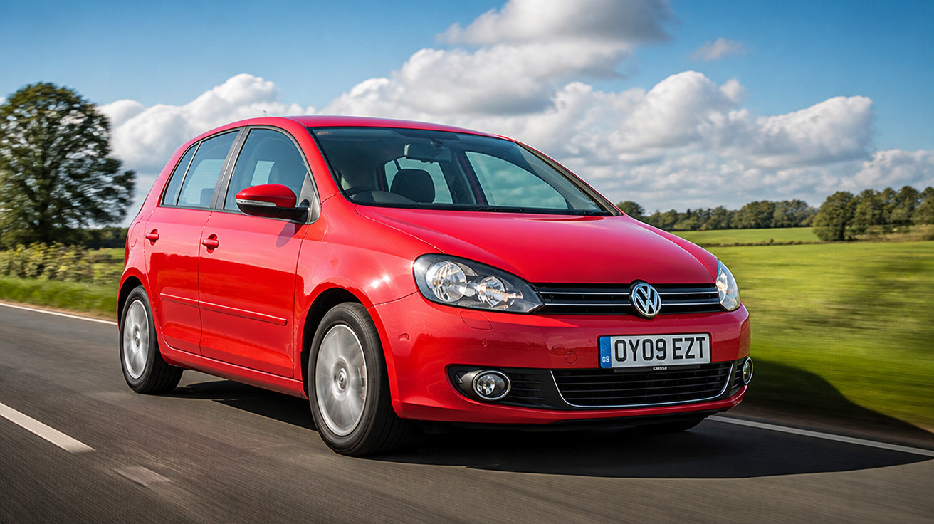 Carro hatchback vermelho Volkswagen em estrada com céu azul e árvores ao fundo.
