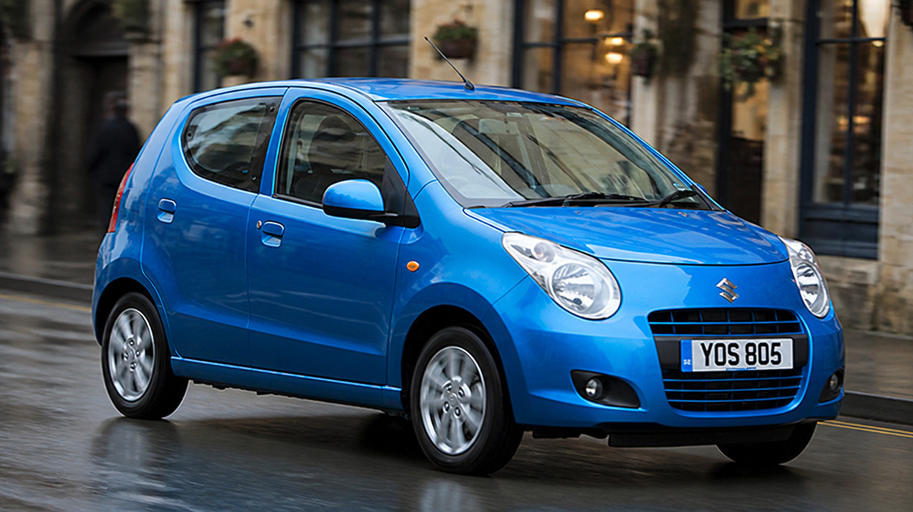 Carro hatch compacto azul Suzuki em rua molhada, com prédios antigos ao fundo.