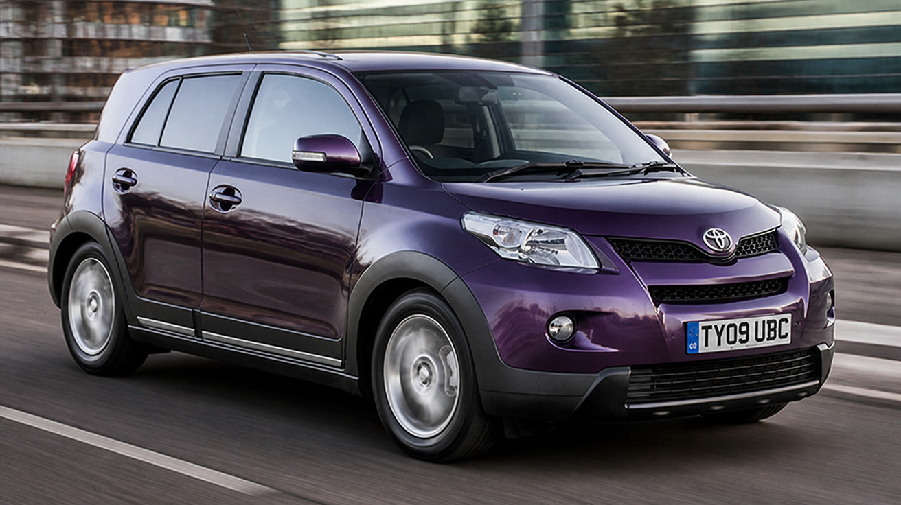 Carro compacto roxo da marca Toyota trafegando em via urbana com prédios ao fundo.