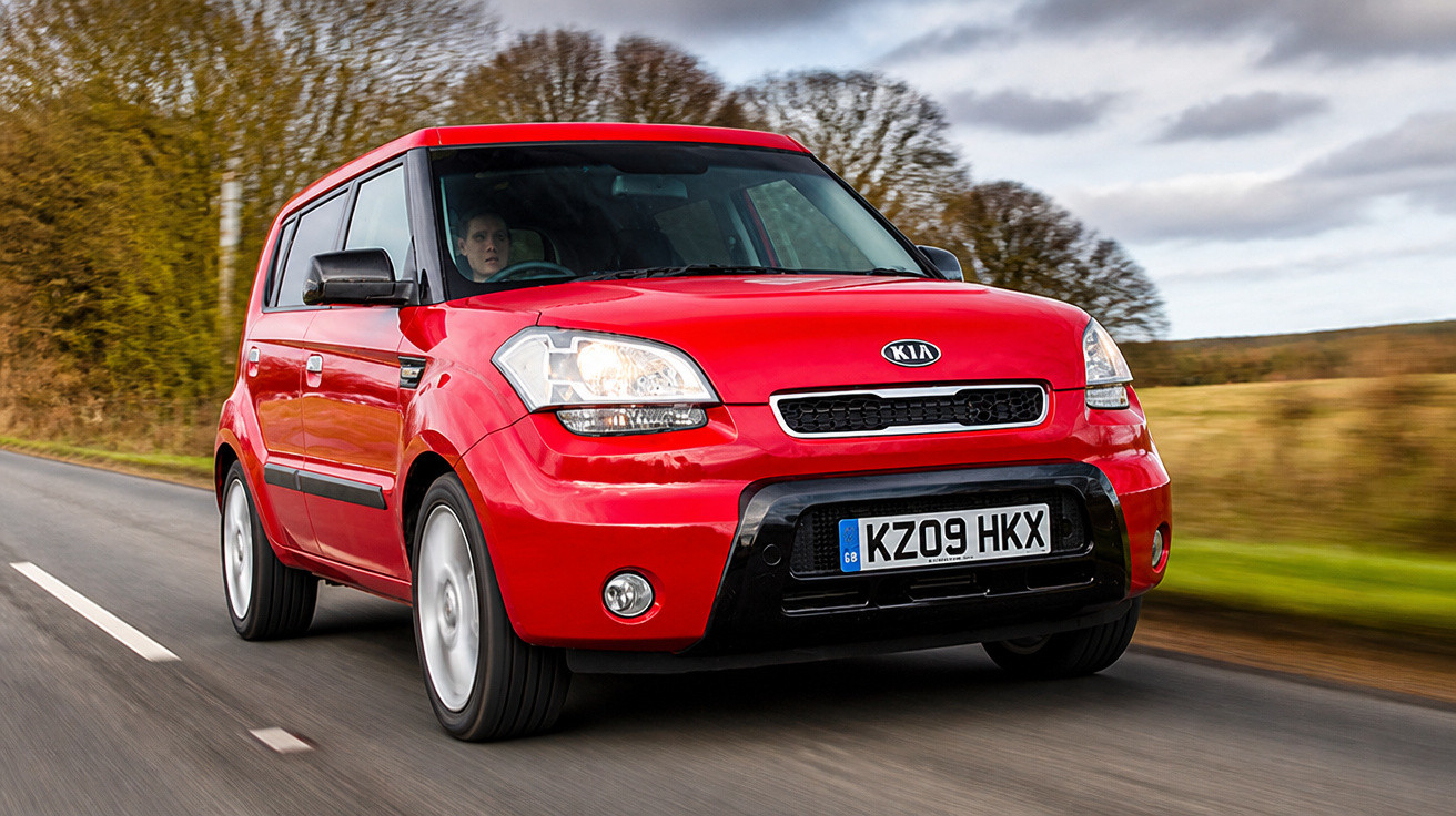 Carro vermelho Kia Soul em movimento na estrada com paisagem de árvores e céu nublado ao fundo.
