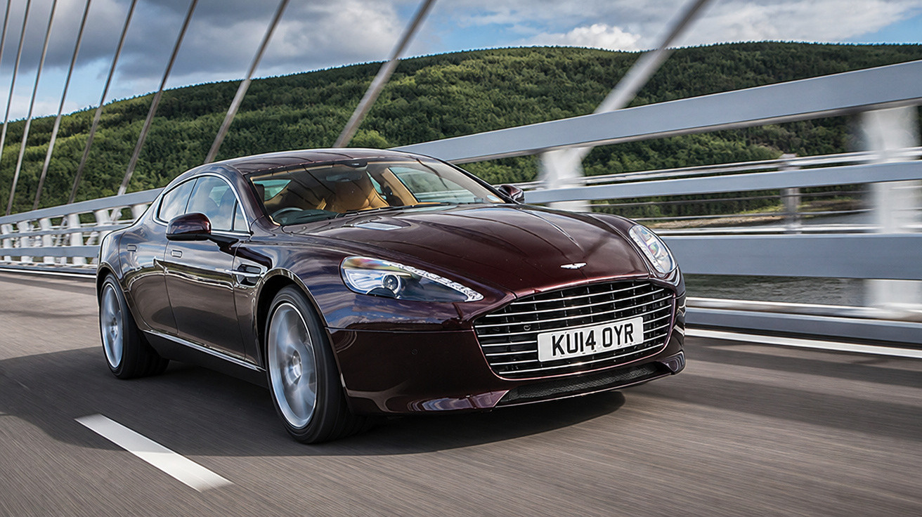 Carro esportivo Aston Martin marrom escuro em movimento em ponte com vegetação ao fundo.
